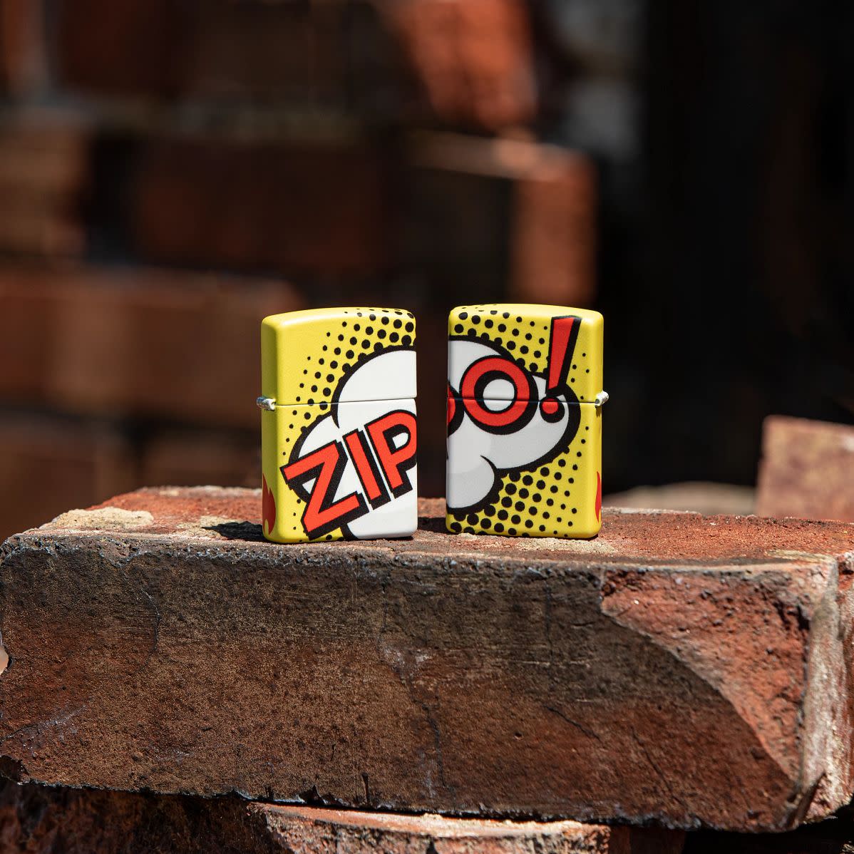 Lifestyle image of two Zippo Pop Art Design 540 Color Windproof Lighters standing on a brick, with one lighter showing the front and the other showing the back